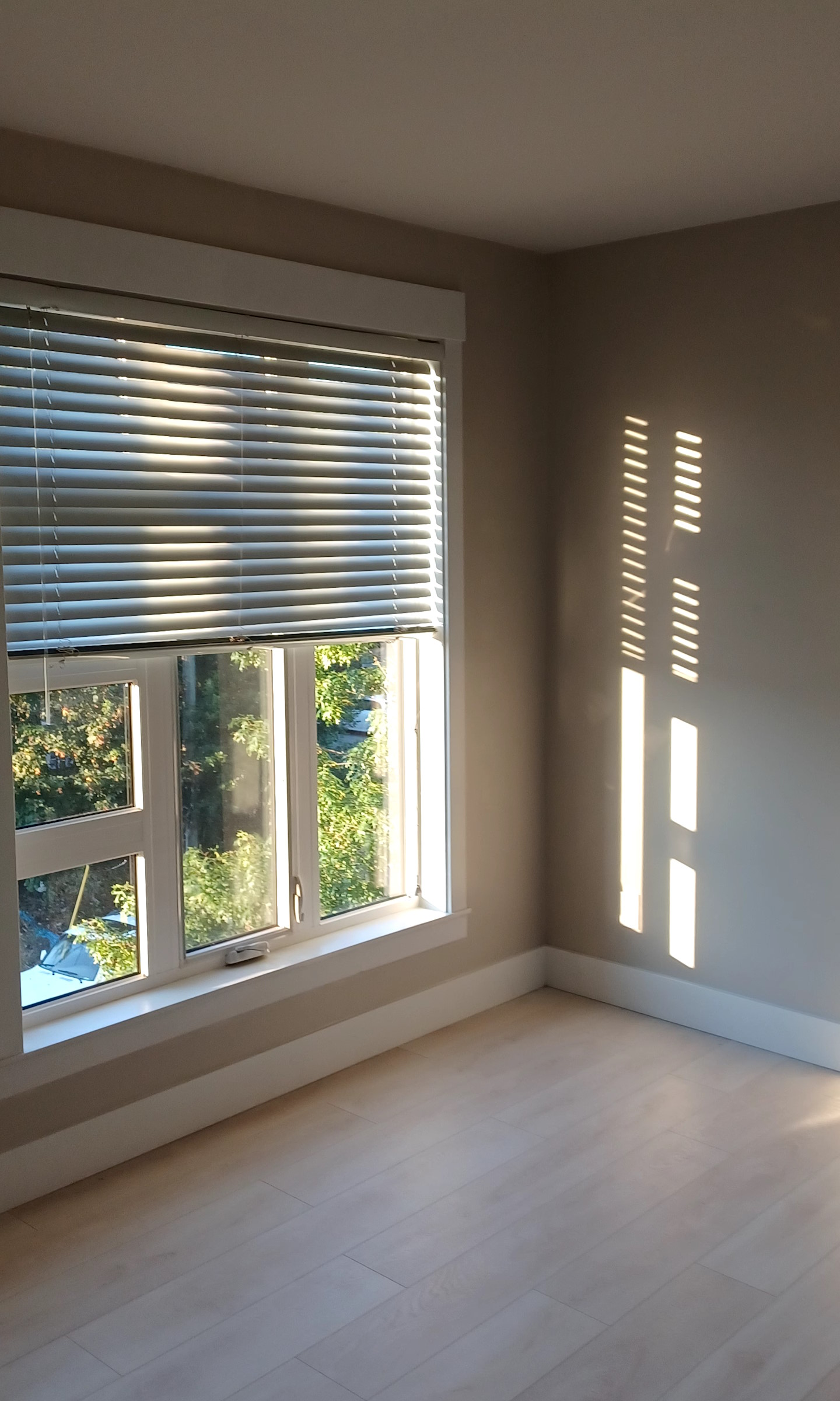 A newly-installed window, with sunlight beaming through the blinds and against a newly-painted light brown wall.